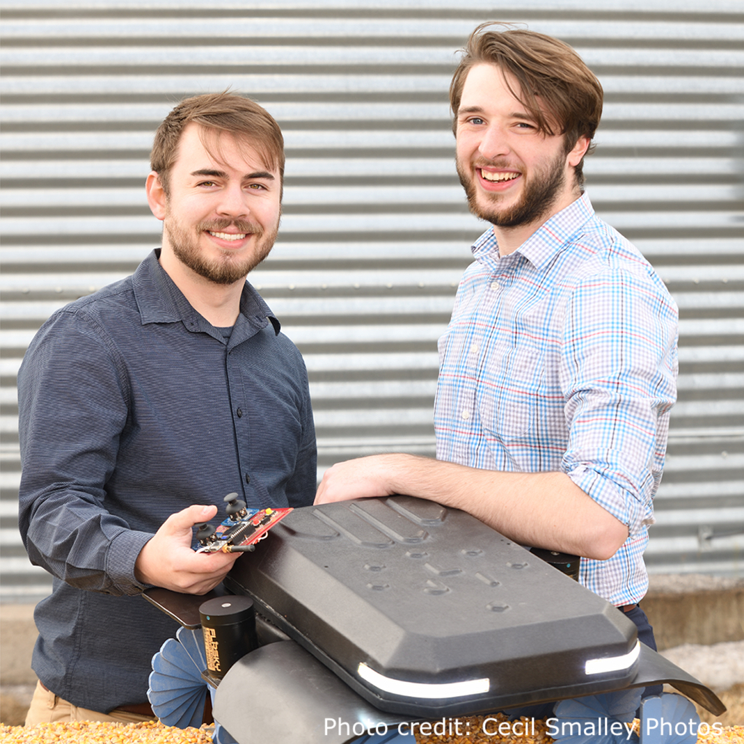 University of Nebraska Omaha undergraduate students Benjamin Johnson and Zane Zents with their Grain Weevil robot that is sitting on top of a pile of grain.